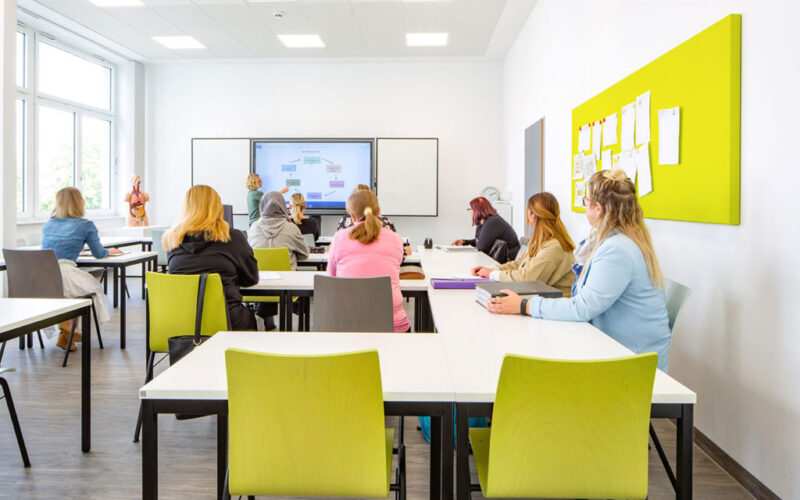 Zu sehen ist ein Klassenraum der Pflegeakademie. Auszubildende sitzen verteilt im Raum und schauen in Richtung der Lehrerein, die an einer digitalen Tafel steht.